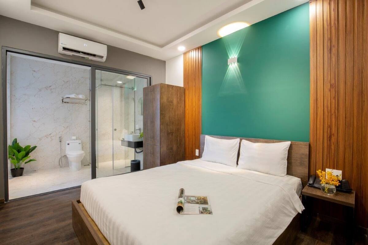 A modern hotel room with a large bed, a wooden headboard, a green accent wall, a glass shower enclosure, and a small bedside table with a vase of flowers.
