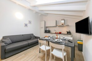 Una sala de estar moderna con un sofá gris, una mesa de comedor preparada para cuatro comensales, y una zona de cocina con un televisor de pantalla plana.