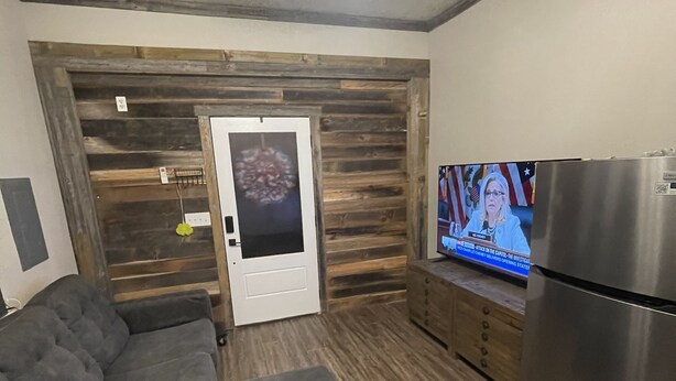 A modern living room with a sectional sofa, a wooden accent wall, a door with a glass panel, a stainless steel refrigerator, and a television displaying a news broadcast.