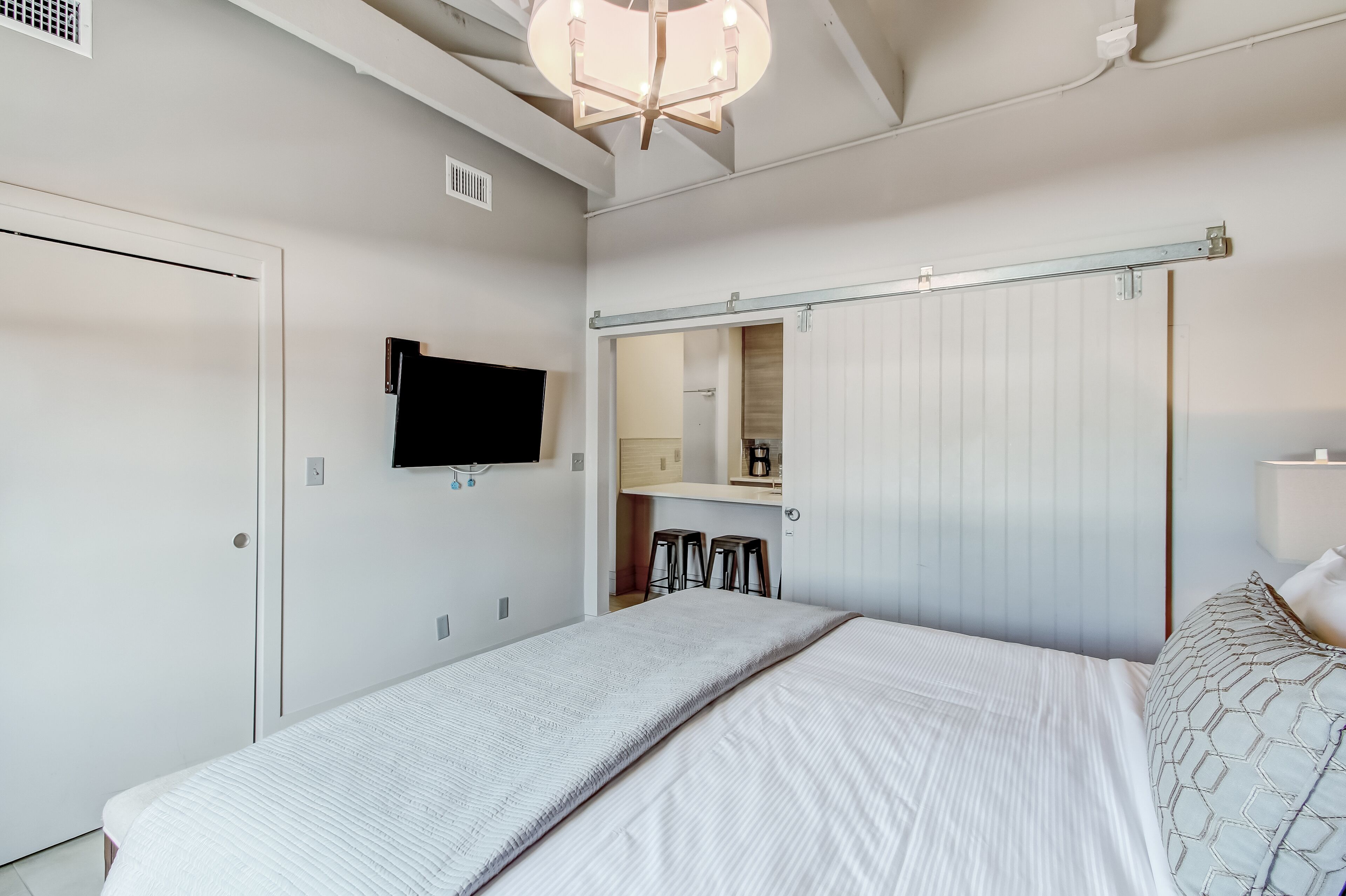 Un dormitorio con cama, televisor montado en la pared, puerta corrediza y una pequeña cocina con taburetes.