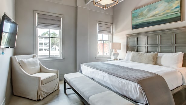 A bedroom with a large bed, a bench, a chair, a TV, and two windows. - Modern Interior, Heated Pool Access (Savannah)