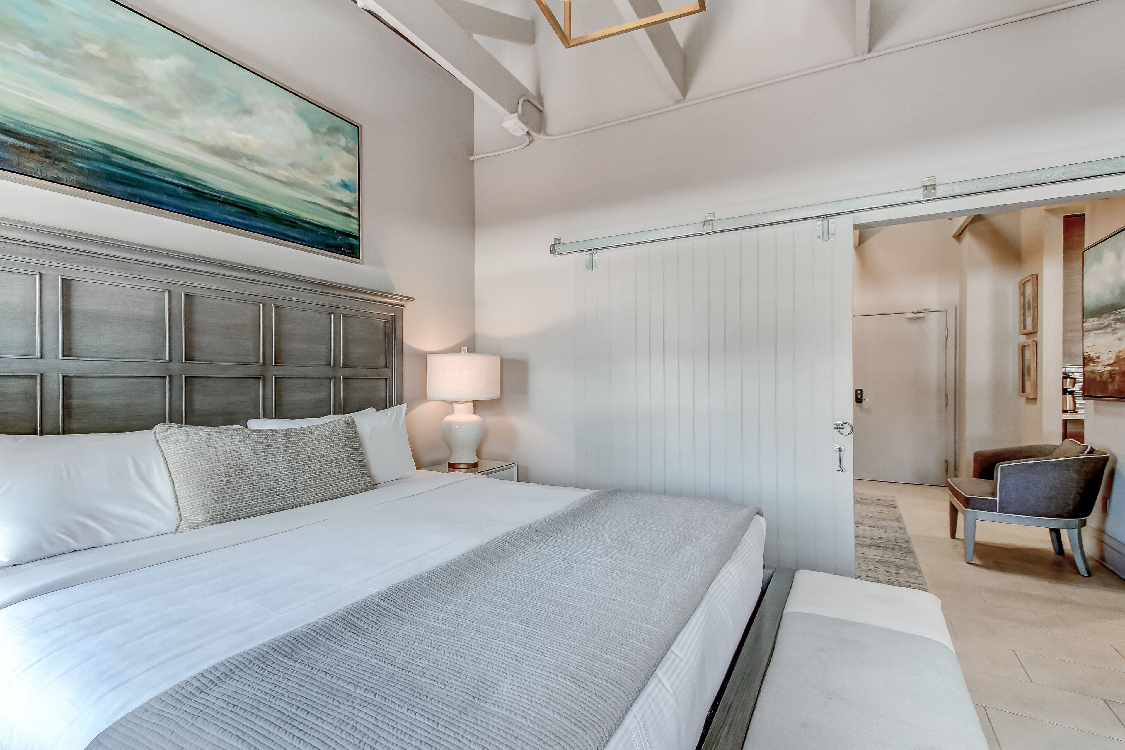 Moderni hotellihuone, jossa on suuri sänky, yöpöytä lampun kanssa, näkymä käytävään ja taulu seinällä.