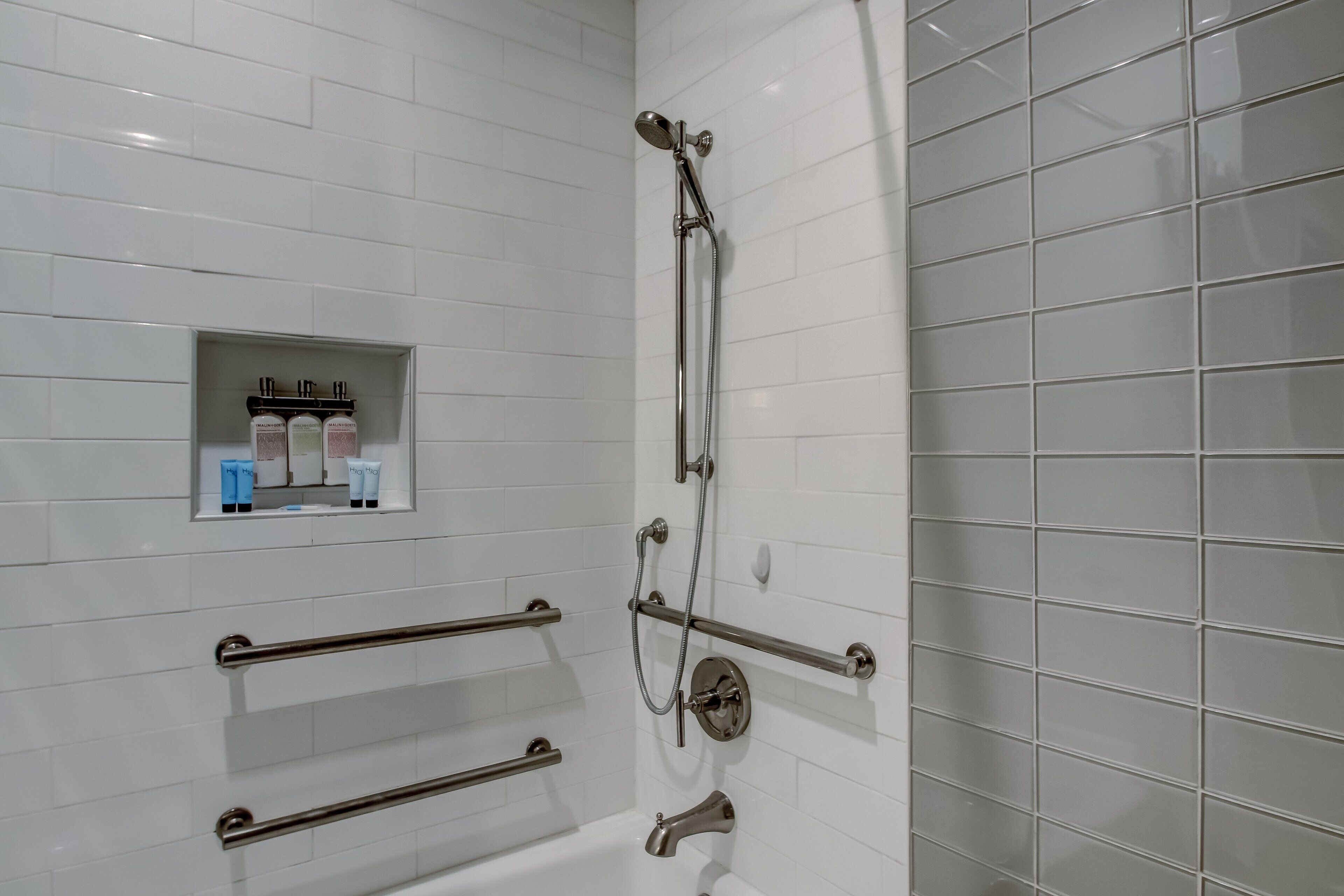 A bathroom with white tiled walls, a shower with a handheld showerhead, and a shelf with toiletries.