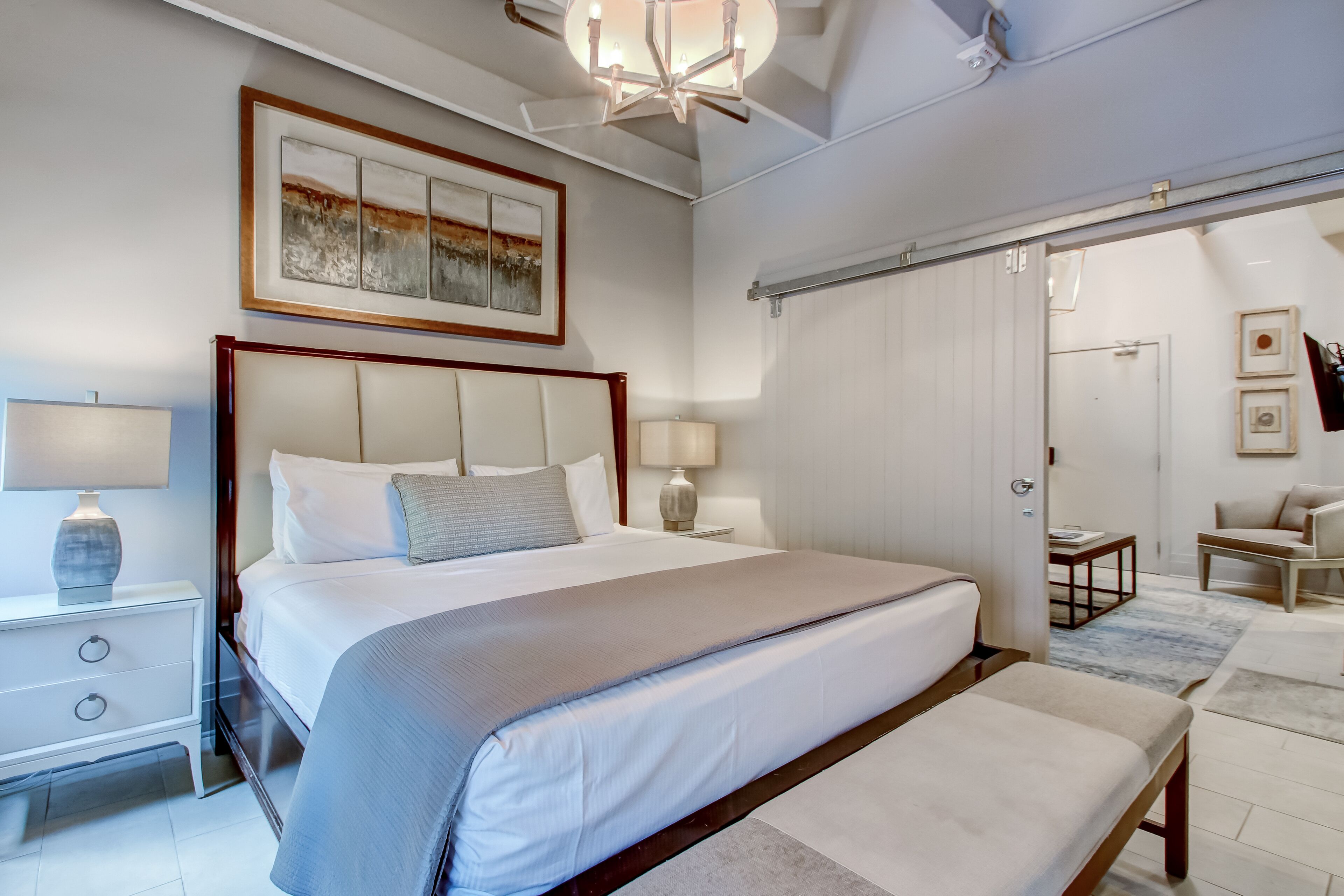 Quarto moderno com uma cama grande, mesas de cabeceira, um candelabro e uma porta deslizante que dá para outro cômodo.