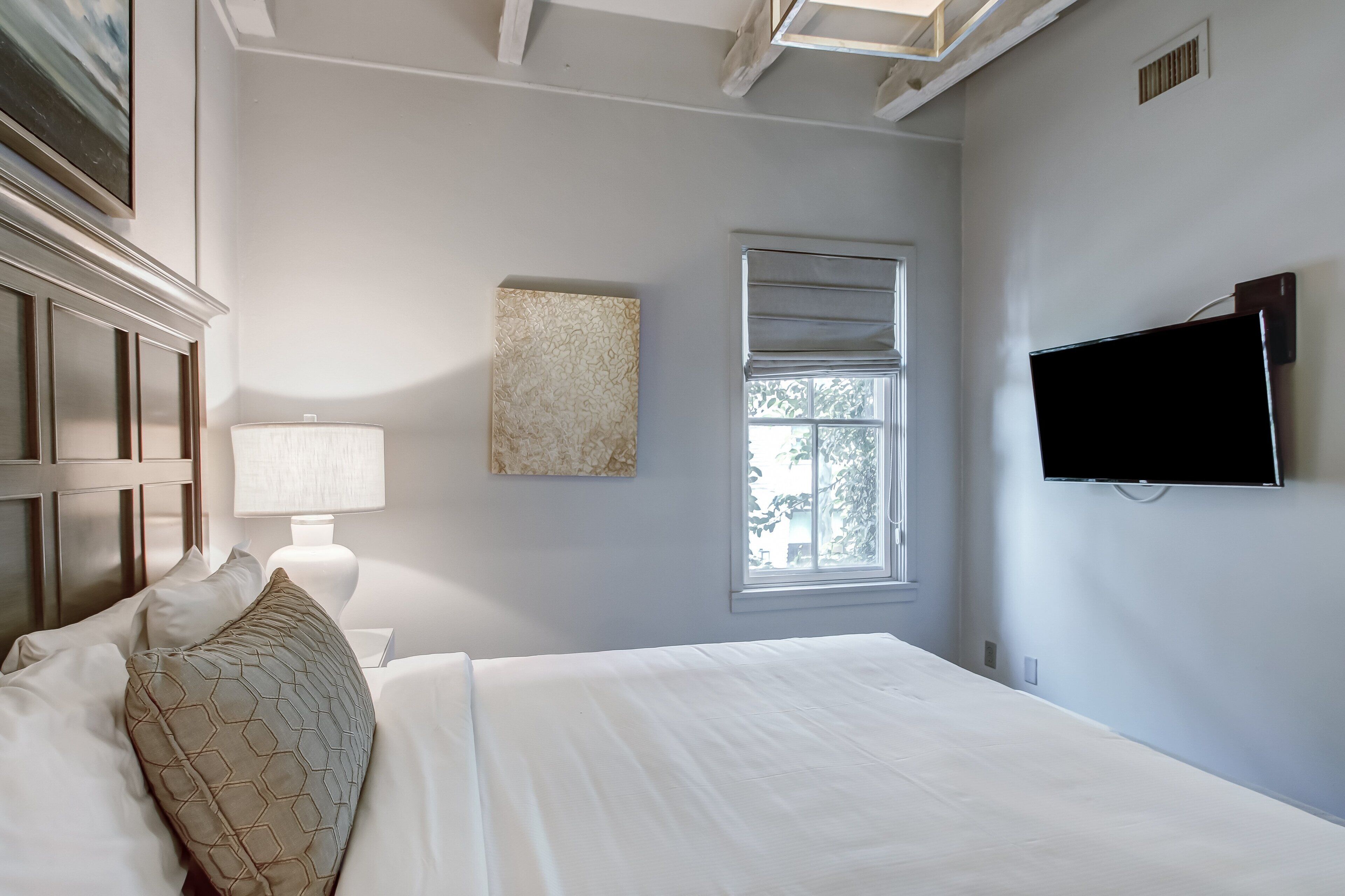 Une chambre à coucher avec un lit, une table de chevet, une lampe, un tableau, une fenêtre et un téléviseur.
