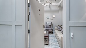 Un salon moderne avec un canapé, un fauteuil et une table basse. Un luminaire au design géométrique est suspendu au plafond, et on aperçoit une porte portant le numéro 7.