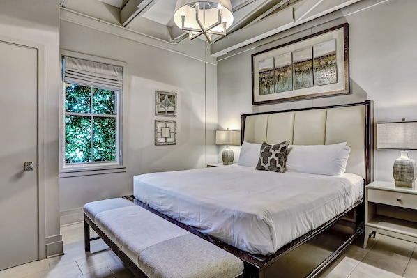 A neatly arranged bedroom with a large bed, a bench, a bedside table with a lamp, and a window with blinds.