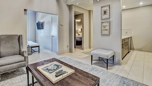 A modern living room with a sofa, a coffee table with remote controls, a bench, and a hallway leading to a bedroom. - Modern Interior, Heated Pool Access (Savannah)