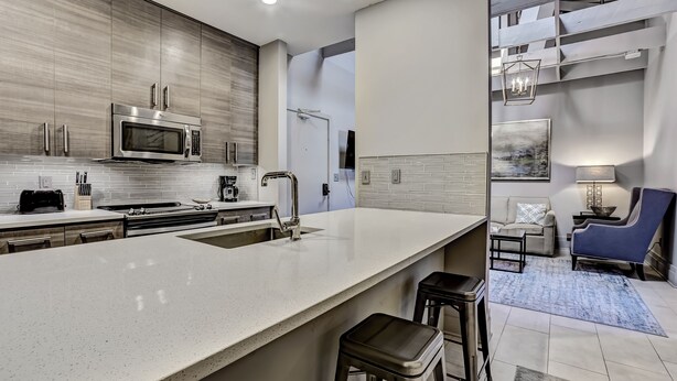 A modern kitchen with a large island, stainless steel appliances, and a dining area with a blue chair and a sofa. - Modern Interior, Heated Pool Access (Savannah)