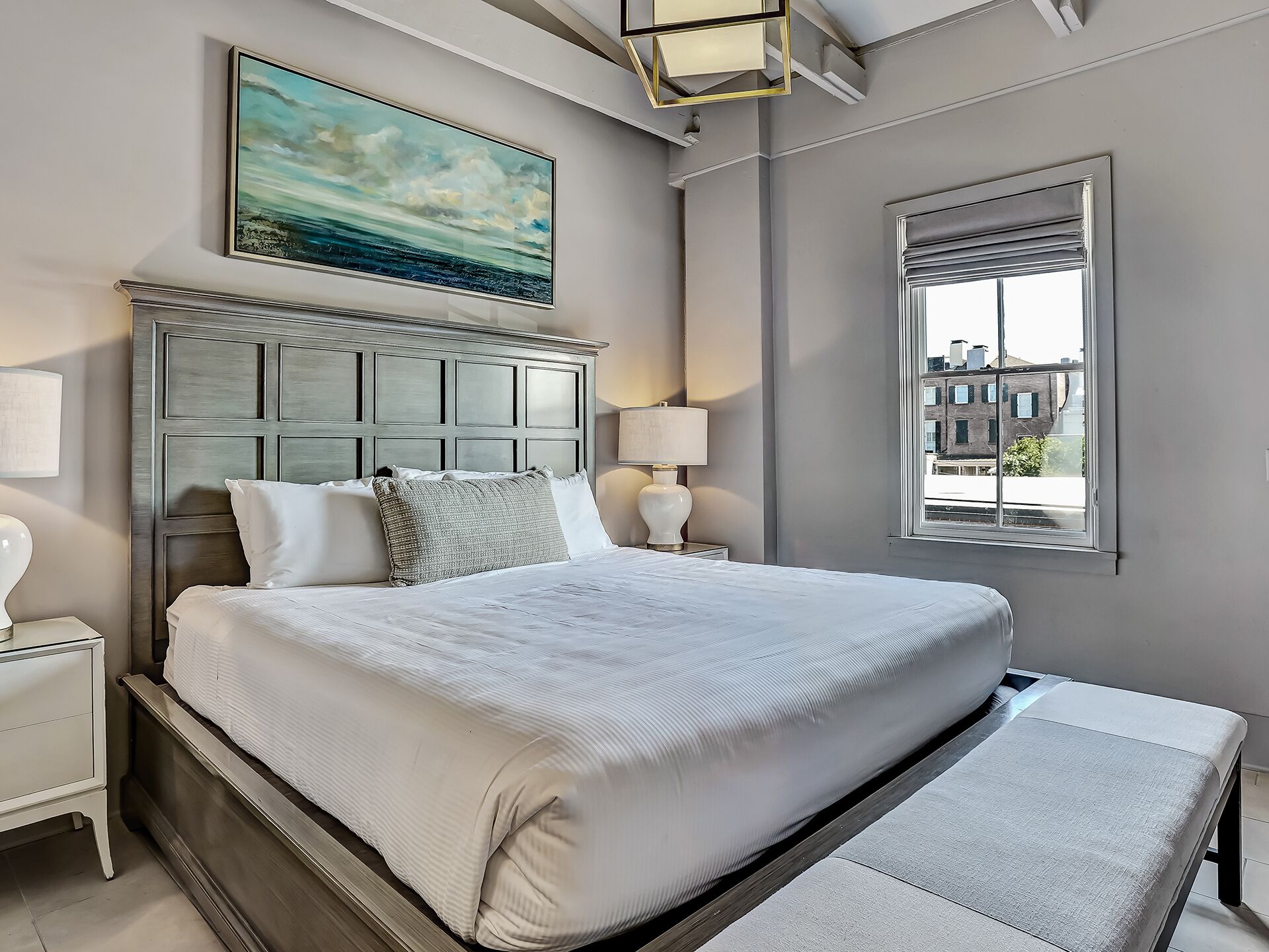A neatly made bed with a headboard, a bedside lamp, and a window with a view of buildings.