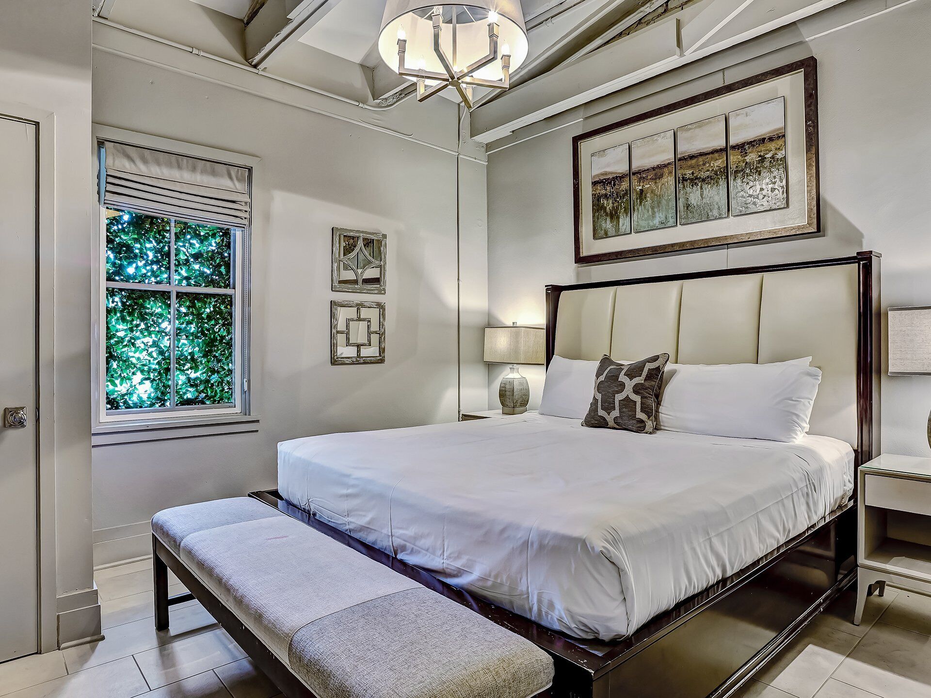 A neatly arranged bedroom with a large bed, a bench, a bedside table, and a window with a view of greenery.