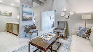 A spacious living area with a modern design, featuring a glass coffee table, two armchairs, a sofa, and a kitchen area in the background. - Modern Interior, Heated Pool Access (Savannah)
