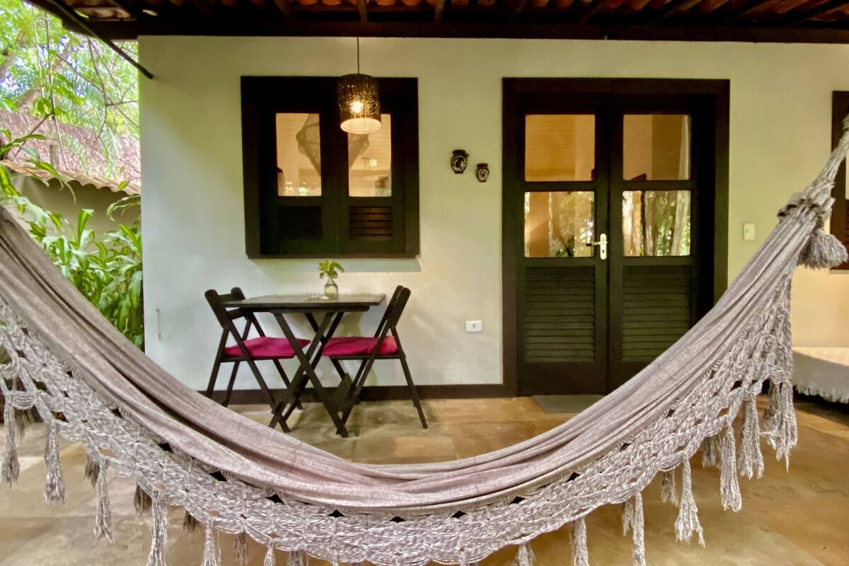 Una hamaca en un espacio al aire libre techado, con una mesa, sillas, una puerta y una ventana con contraventanas.