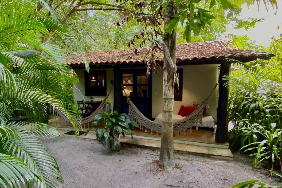 Una casita con techo de tejas, una hamaca y una cama con almohada roja.