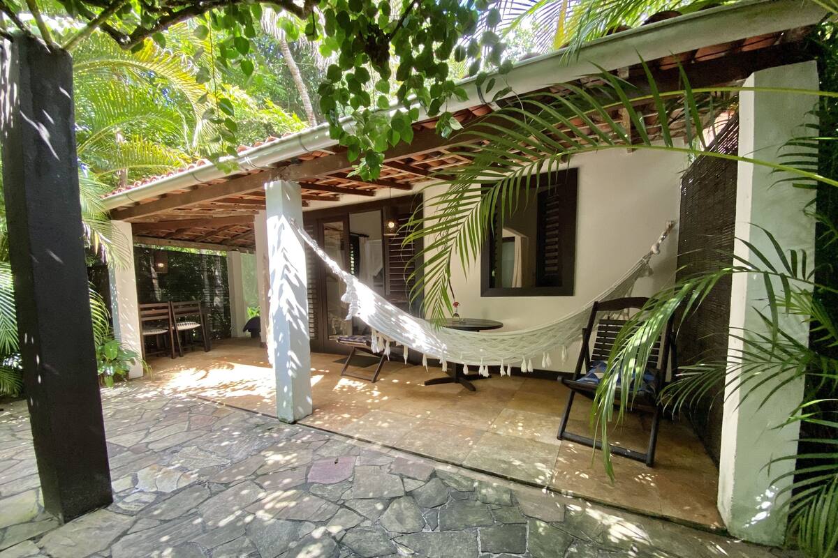Un patio techado con una hamaca, suelo de piedra y abundante vegetación.