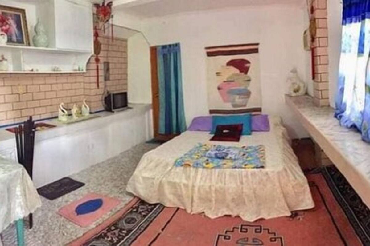 A bedroom with a bed, a small table, a chair, a window with curtains, and a brick wall.