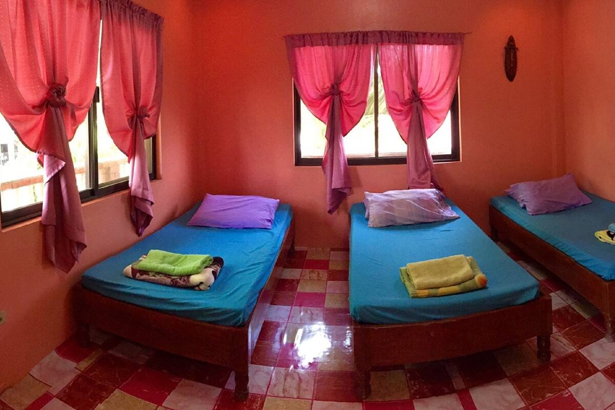 A room with three single beds, each with a blue mattress and a pillow. The room has orange walls and a checkered floor.