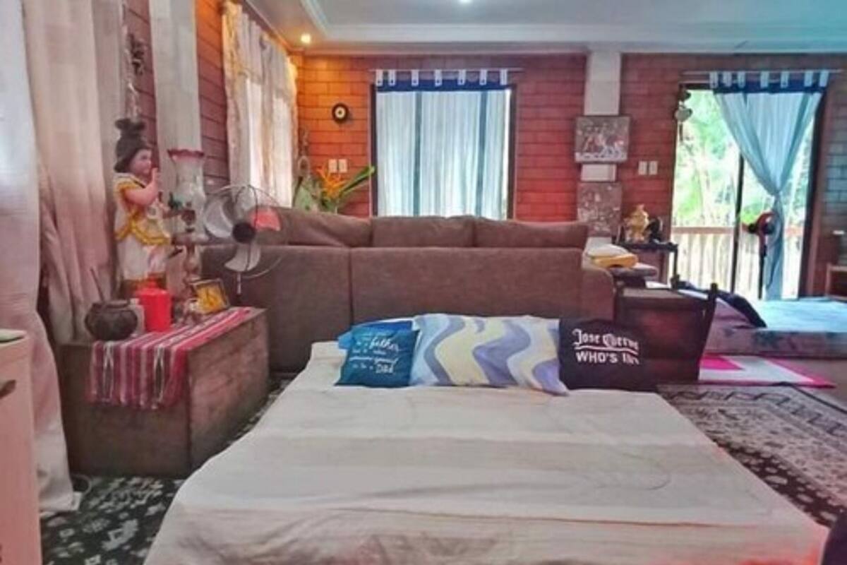 A room with a bed, a sofa, a small table with a fan, and a window with curtains.