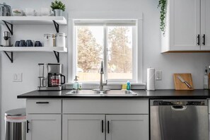 Une cuisine moderne Ă©quipĂ©e dâun lave-vaisselle en acier inoxydable, dâun Ă©vier Ă deux bacs et dâune fenĂȘtre donnant sur les arbres.
