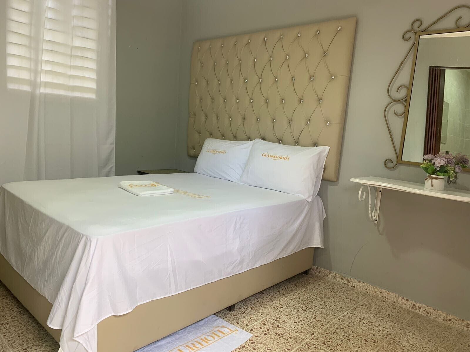 A neatly made bed with a tufted headboard, a bedside table with a mirror, and a small vase with flowers.