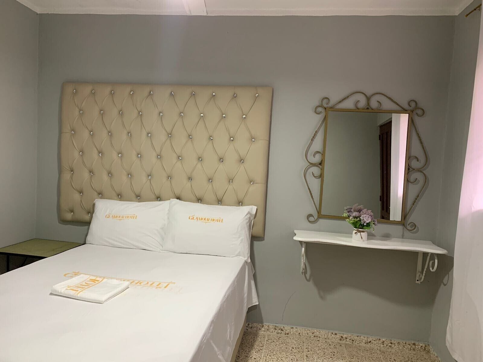 A bedroom with a tufted headboard, a bed with white linens, a bedside table with a mirror, and a small vase with flowers.
