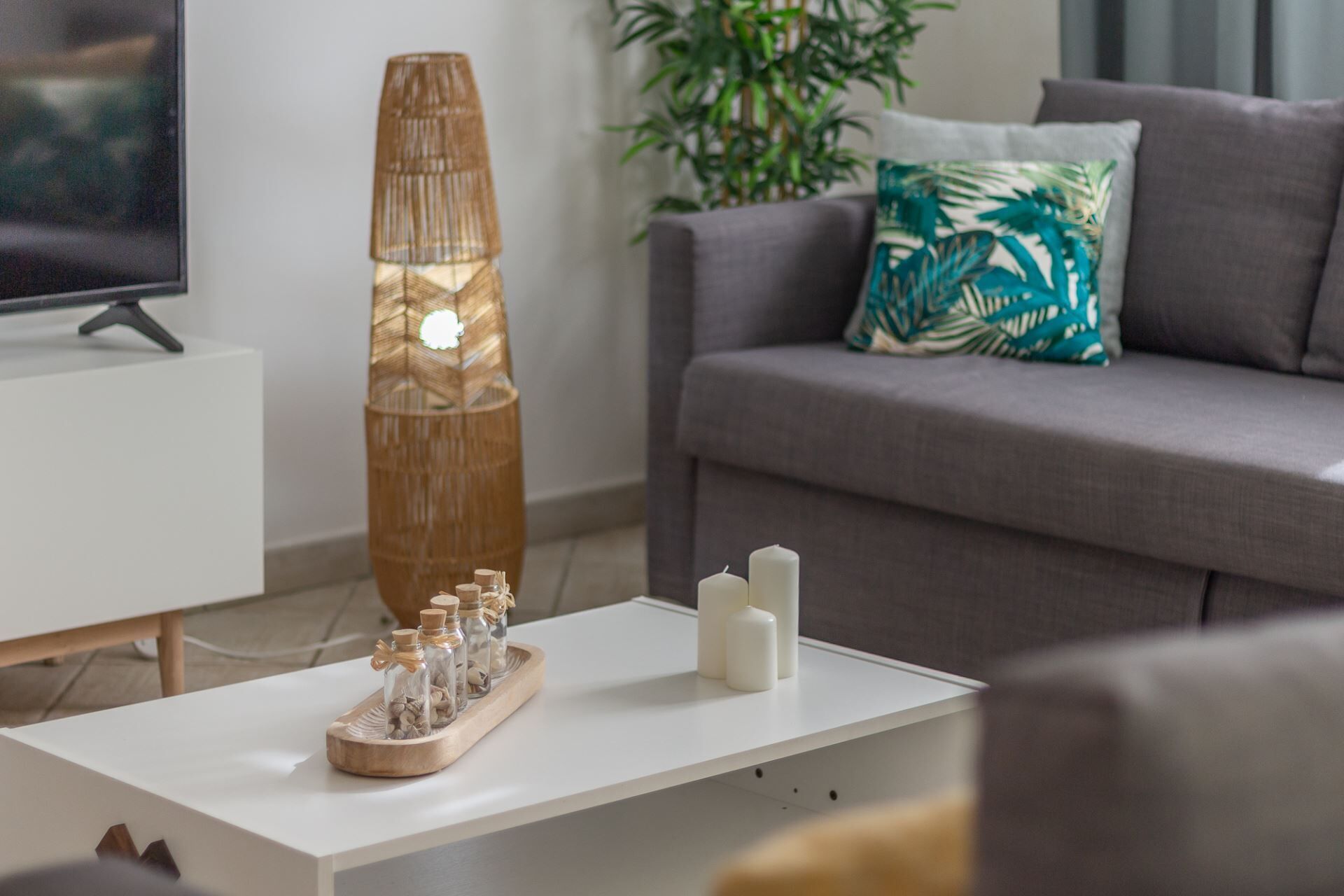 A modern living room with a white coffee table, a tall bamboo lamp, and a grey sofa with a tropical leaf pillow.