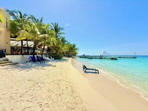 A beach with clear turquoise water, palm trees, and a pier with boats. There are lounge chairs and a building with a covered area.