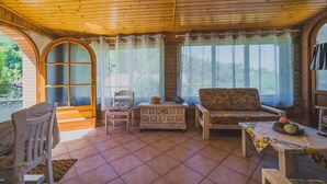 A cozy room with wooden ceiling and tiled floor, featuring a sofa, a chair, a small table with a basket, and a window with curtains. - UN GIARDINO SU PARETI (Capoliveri)