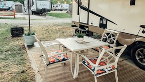 Een camping met een tafel en stoelen onder een overkapping, campers op de achtergrond, en een bordje met de tekst 'WILDWOOD HERITAGE GLEN'.