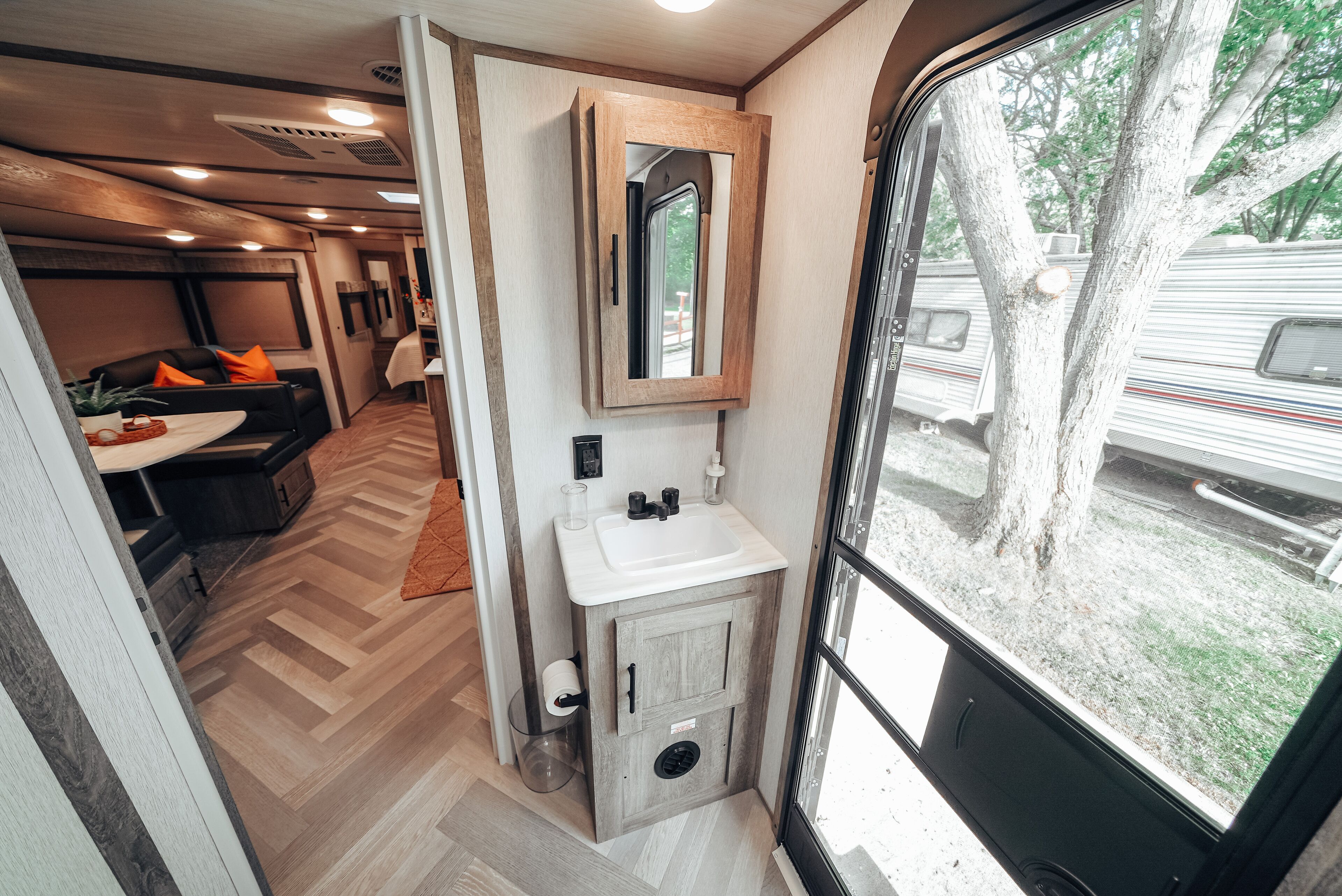 A compact RV interior with a kitchenette, a small seating area, and a window with a view of an RV park.