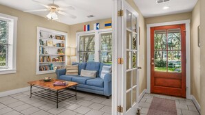 A living room with a blue sofa, a coffee table, and a bookshelf. There is a door with glass panels and a red doorbell. - Family Paradise on Lands End - Private Dock (Saint Helena Island)