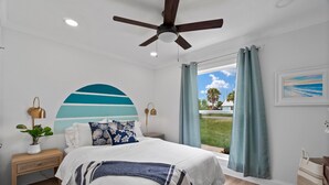 Ein Schlafzimmer mit Bett, Deckenventilator, Nachttisch, Lampe, Pflanze und einem Fenster mit Vorhängen.