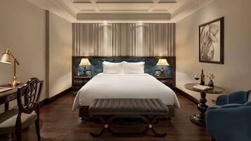 A hotel room with a large bed, two bedside tables with lamps, a chair, a round table with a vase, and a framed artwork on the wall.