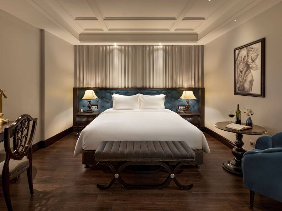 Quarto de hotel com uma cama grande, duas mesas de cabeceira com luminárias, uma cadeira, uma mesa redonda com um vaso e uma obra de arte emoldurada na parede.