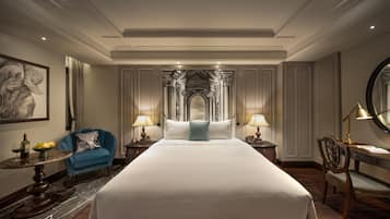 Habitación de hotel con una cama grande, dos mesitas de noche con lámparas, un sillón azul, una mesa redonda con una botella y fruta, y un cuadro en la pared.