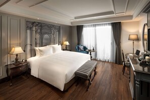 A hotel room with a large bed, a desk with a chair, a small table with a lamp, and a large mirror.