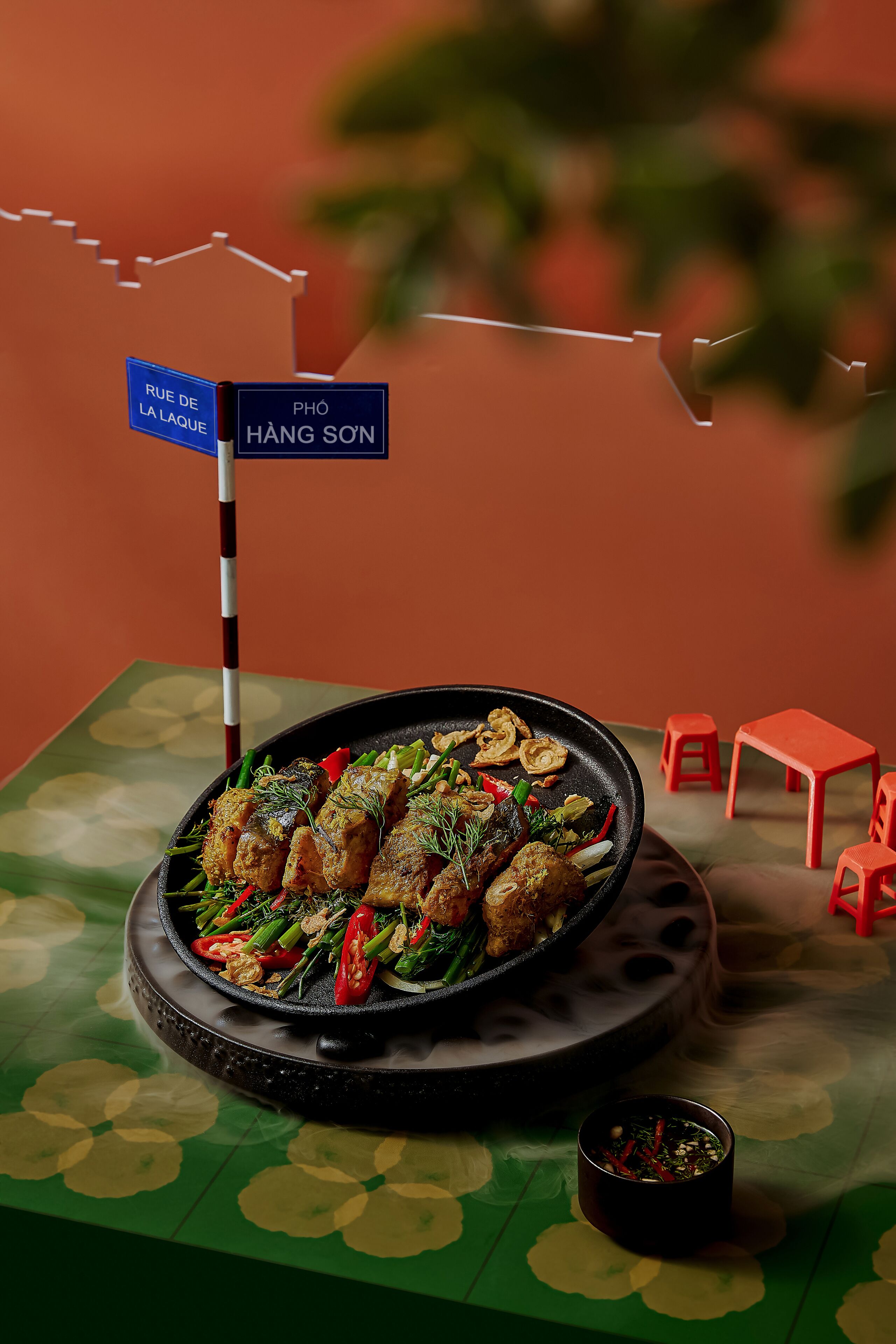 Une table à manger avec une assiette noire contenant des aliments, un petit bol de sauce et une enseigne indiquant « RUE DE LA LAQUE » et « PHO HÀNG SẺN ».