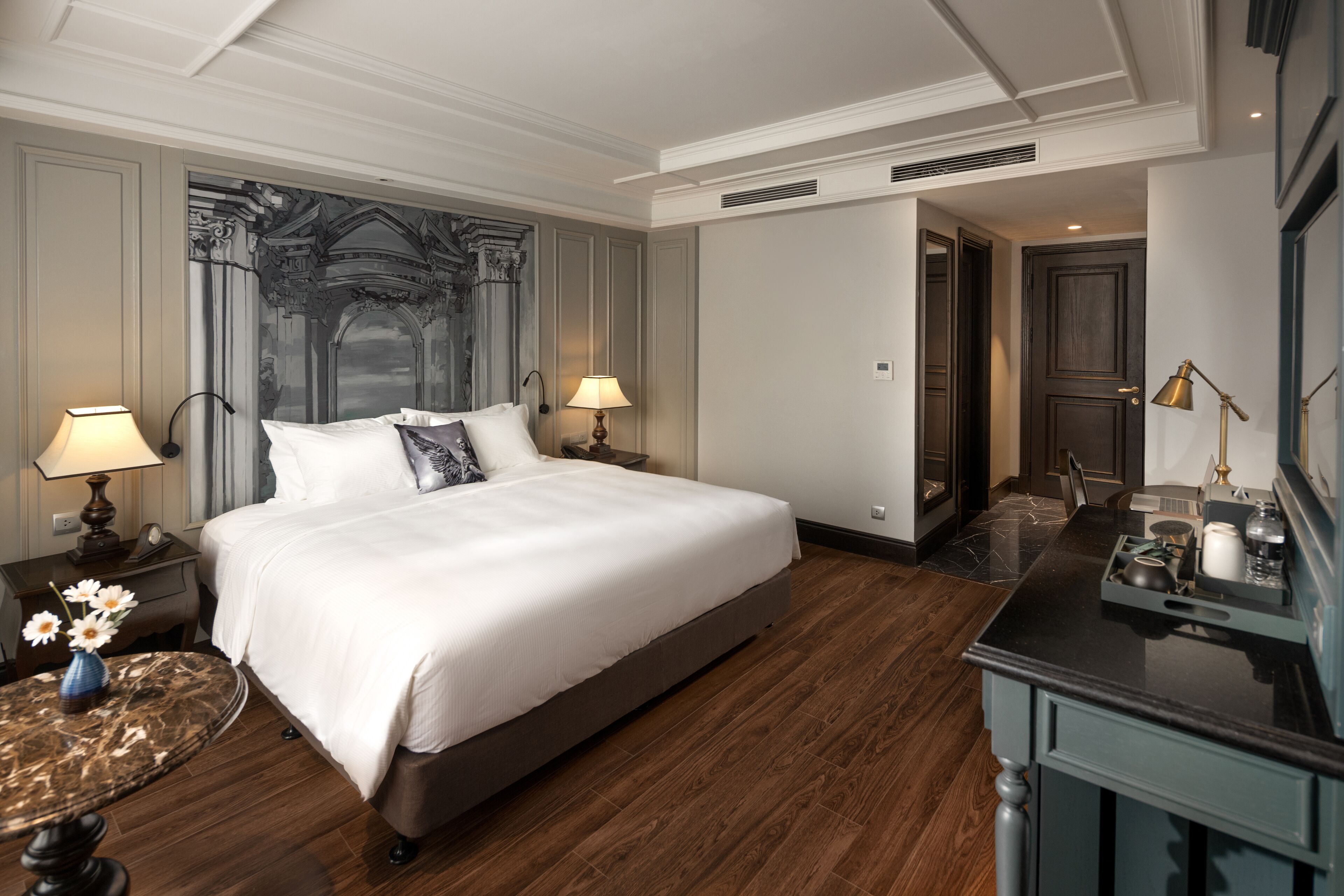 Une chambre d’hôtel avec un grand lit, deux tables de chevet avec des lampes, une petite table ronde avec un vase et un parquet en bois foncé.