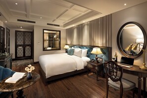 A hotel room with a large bed, a desk with a laptop, a chair, a round mirror, and a small table with a vase of flowers.
