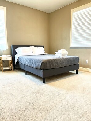 Una cama bien hecha en una habitación con paredes color beige, una mesita de noche pequeña y una ventana con persianas.