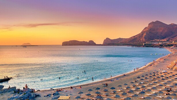 Une plage avec des parasols et des chaises longues, un ciel dégagé et des montagnes en arrière-plan.