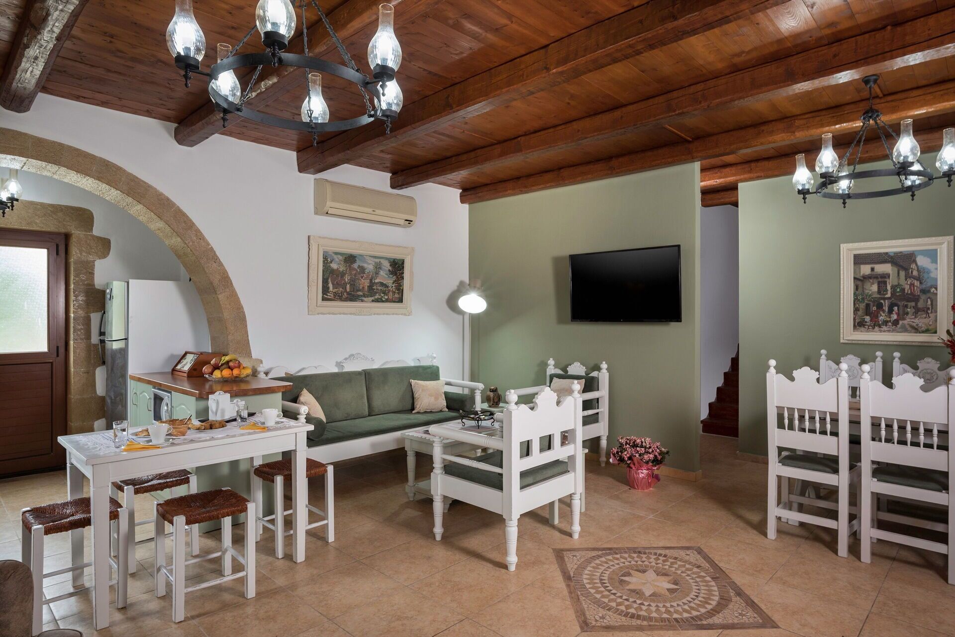 A cozy living room with a wooden ceiling, a green sofa, a white coffee table, a white chair, a small table with a fruit bowl, and a TV mounted on the wall.