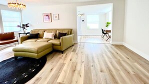 Amplia sala de estar con un sofá seccional grande, una alfombra negra y piso de madera. Se ve un comedor con mesa y sillas al fondo.