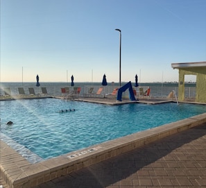 Una piscina con sillas de descanso y sombrillas, un puesto de socorrista y una vista despejada del océano.