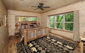 A wooden cabin bedroom with a bed, a nightstand, a ceiling fan, and a large window with a view of trees. - Swim Year-Round, Sip Fireside, Host Movie Nights, Play Pool & Soak in the Hot Tub - Mystic Timber (Sevierville)