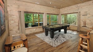 Een pooltafel in een houten kamer met grote ramen en uitzicht op bomen.