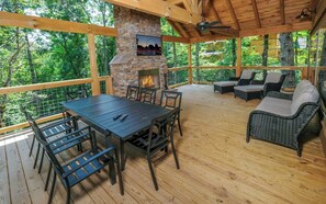 An outdoor deck with a stone fireplace, wooden furniture, and a view of the surrounding forest. - Swim Year-Round, Sip Fireside, Host Movie Nights, Play Pool & Soak in the Hot Tub - Mystic Timber (Sevierville)