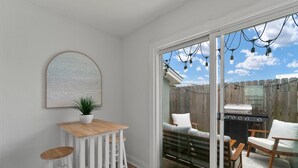 A patio with a wooden table and white bar stools, a grill, and a view of a backyard with a wooden fence. - PCB Modern Beach Casita, Fully Fenced Yard (Panama City Beach)