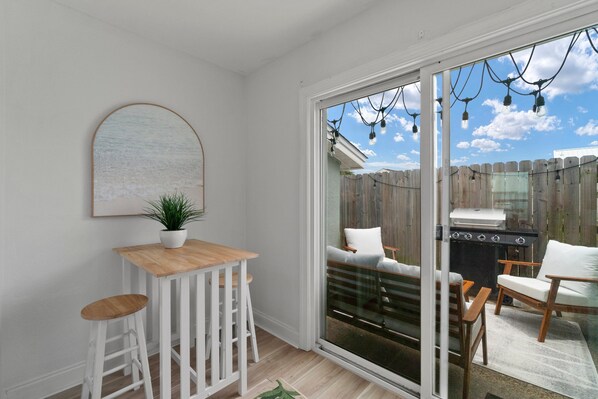 A patio with a wooden table and white bar stools, a grill, and a view of a backyard with a wooden fence.