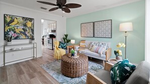 A living room with a round wicker ottoman, a sofa, a coffee table, and a ceiling fan. There are two wall-mounted paintings, a floor lamp, and a potted plant. - PCB Modern Beach Casita, Fully Fenced Yard (Panama City Beach)